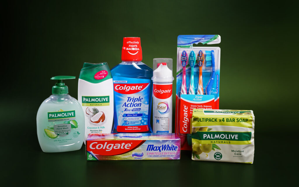 Various Colgate-Palmolive personal care products are lined up next to each other against a dark green background. The Palmolive products include bar soap, liquid soap and shower gel. The Colgate products include toothpaste, mouthwash and toothbrushes.