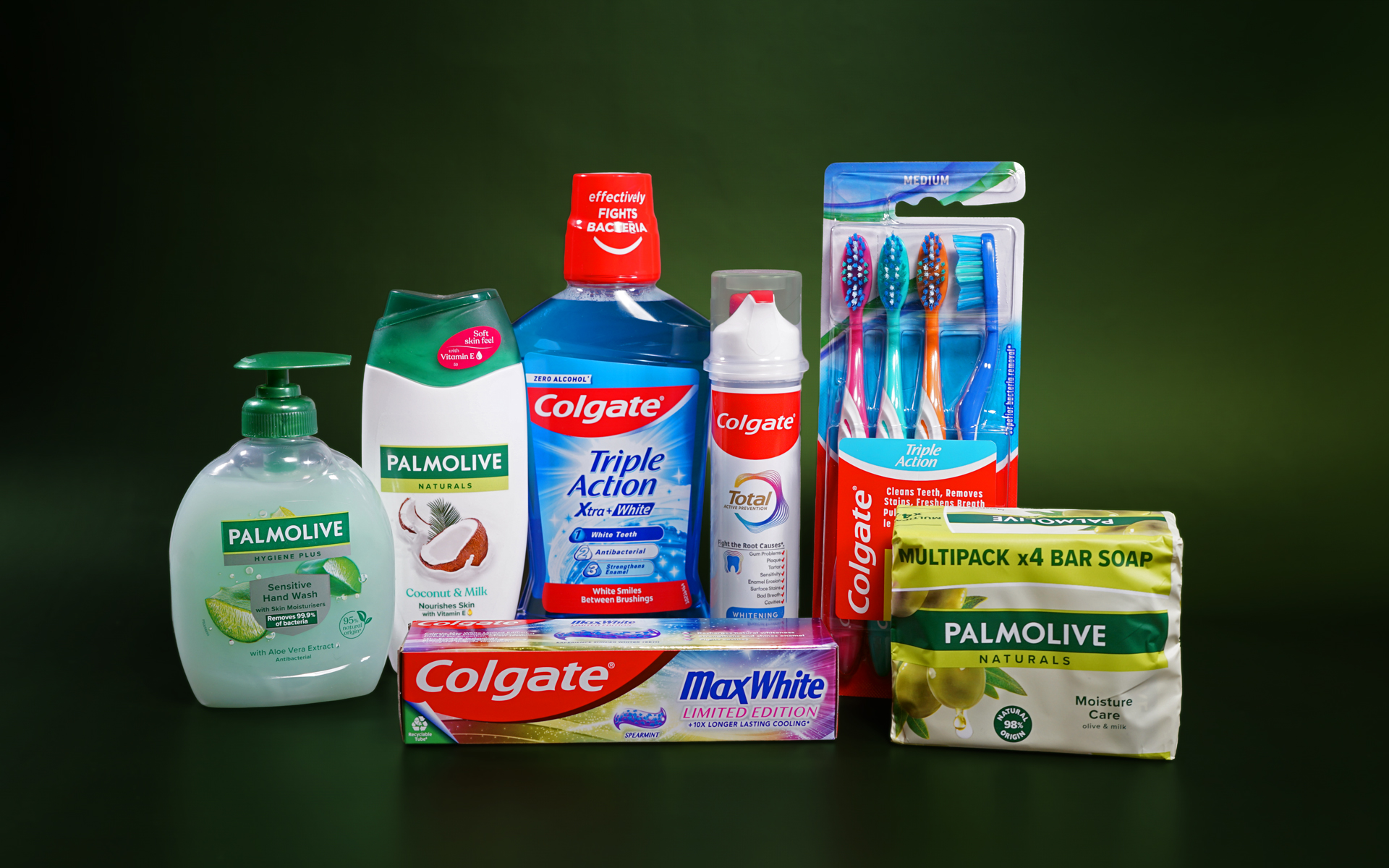 Various Colgate-Palmolive personal care products are lined up next to each other against a dark green background. The Palmolive products include bar soap, liquid soap and shower gel. The Colgate products include toothpaste, mouthwash and toothbrushes.