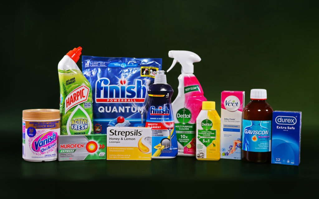 Various cleaning, healthcare and personal care items are lined up next to each other against a dark green background. Some of the items include dishwasher tablets, surface sprays, throat lozenges, pain relief medication, wax strips and condoms. The brands visible are Vanish, Harpic, Nurofen, Strepsils, Finish, Dettol, Veet, Gaviscon and Durex.