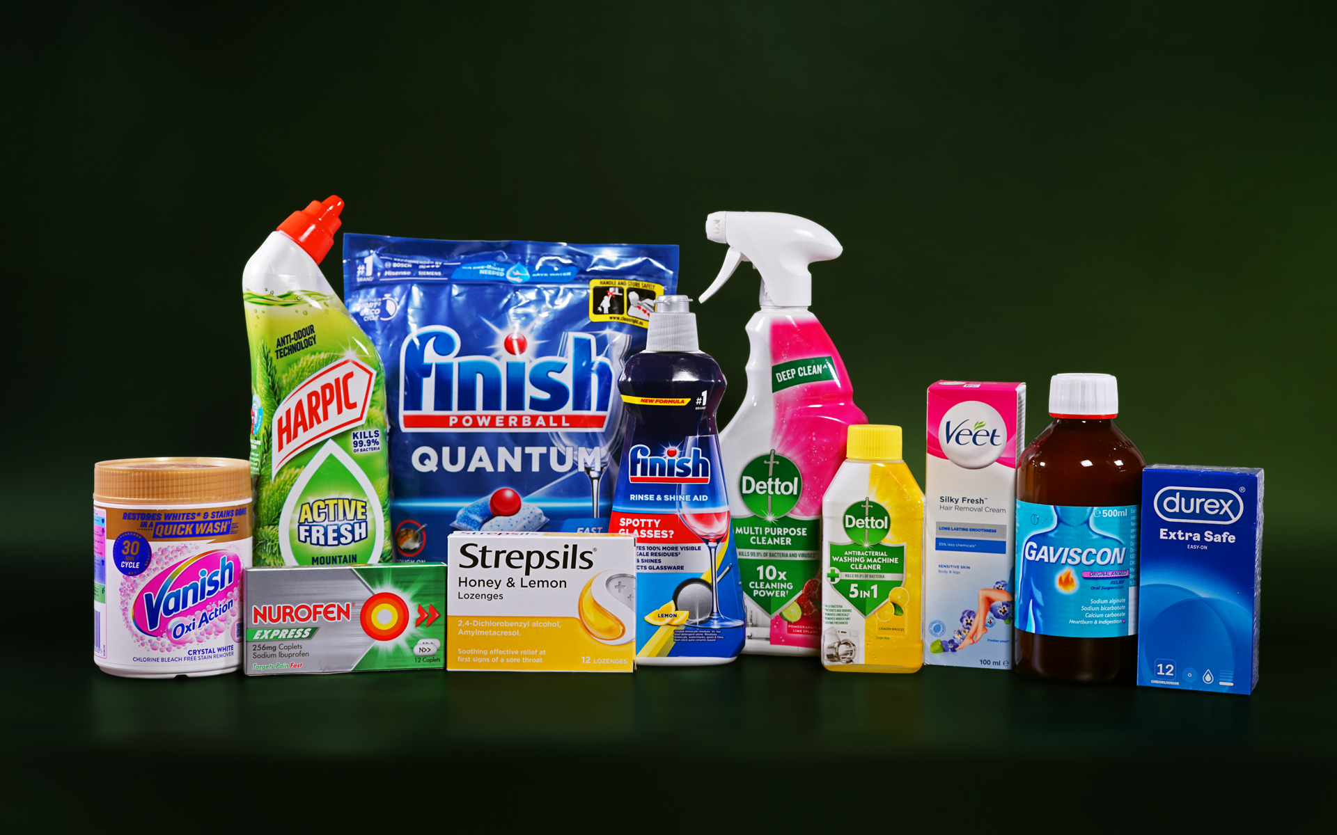 Various cleaning, healthcare and personal care items are lined up next to each other against a dark green background. Some of the items include dishwasher tablets, surface sprays, throat lozenges, pain relief medication, wax strips and condoms. The brands visible are Vanish, Harpic, Nurofen, Strepsils, Finish, Dettol, Veet, Gaviscon and Durex.