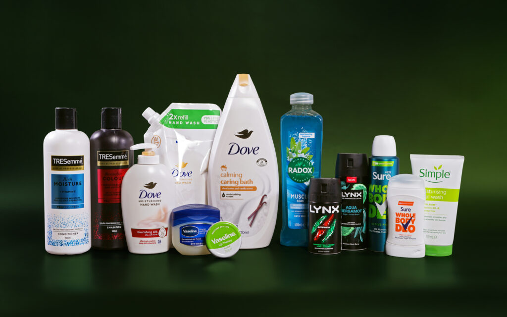 Various personal care products are lined up next to each other against a dark green background. The products include conditioner, shampoo, hand wash, shower gel, petroleum jelly, deodorant and face wash. The brands visible include TRESemme, Dove, Vaseline, Radox, Lynx, Sure and Simple.