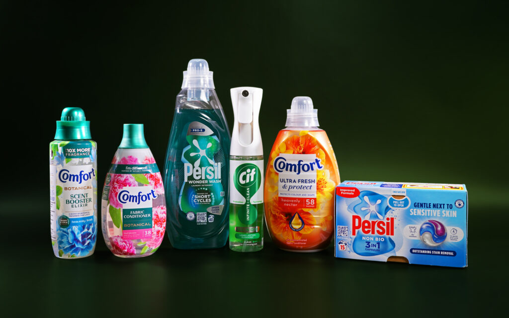 Multiple cleaning products in boxes and bottles are lined up together in a dark green space. The products include Persil liquid laundry detergent, Persil laundry pods, Comfort fabric conditioners, Comfort scent booster and a Cif cleaning spray.