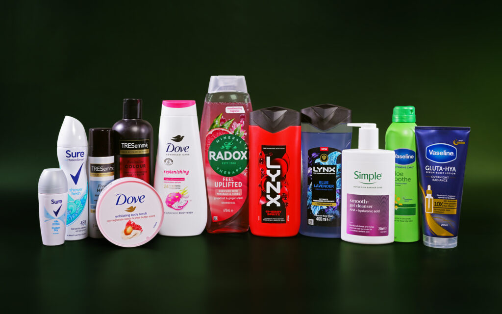Various Unilever Personal Care products are lined up next to each other against a dark green background. These include deodorants, hair products, shower gels and skincare products. The brands visible are Sure, TRESemme, Dove, Radox, Lynx, Simple and Vaseline.
