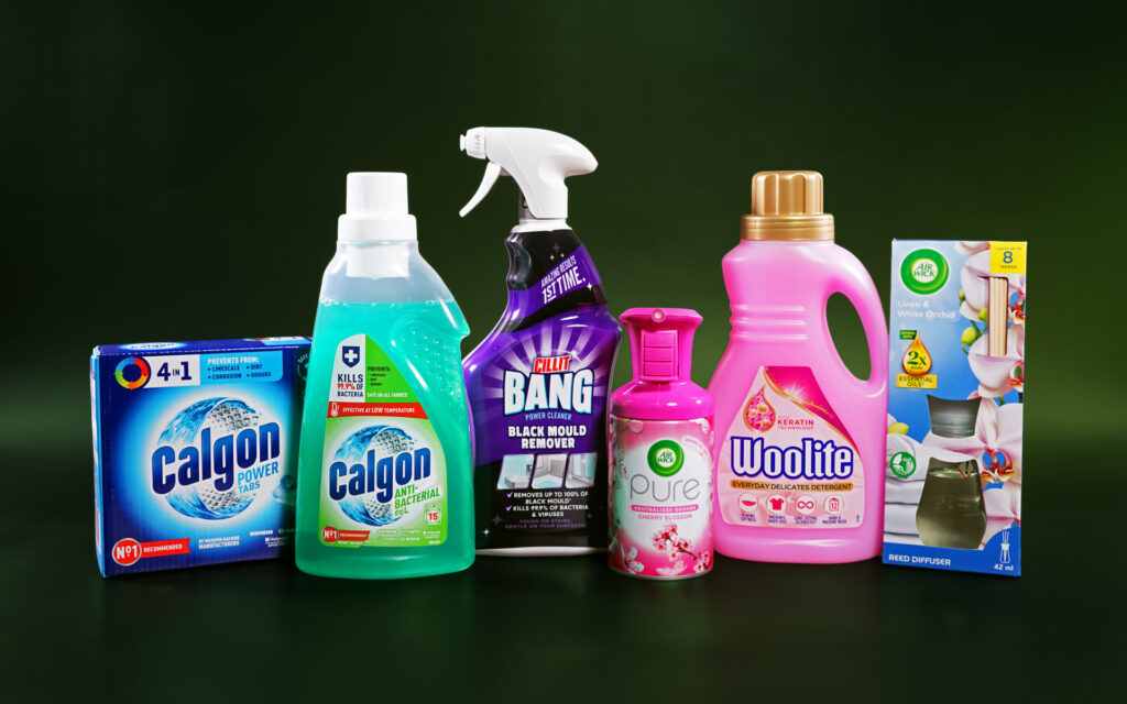 Six home products are lined up next to each other against a dark green background. These products include two Calgon washing machine cleaners, Cillit black mould remover, Woolite laundry detergent and two home scent products from Air Wick.