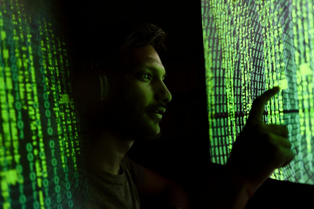 A man is stood in between two green neon text screens in a dark room. His face is lit green with an inquisitive look on his face as he stands facing one of the screens. His hand reaches out to touch one of the screens with his finger.