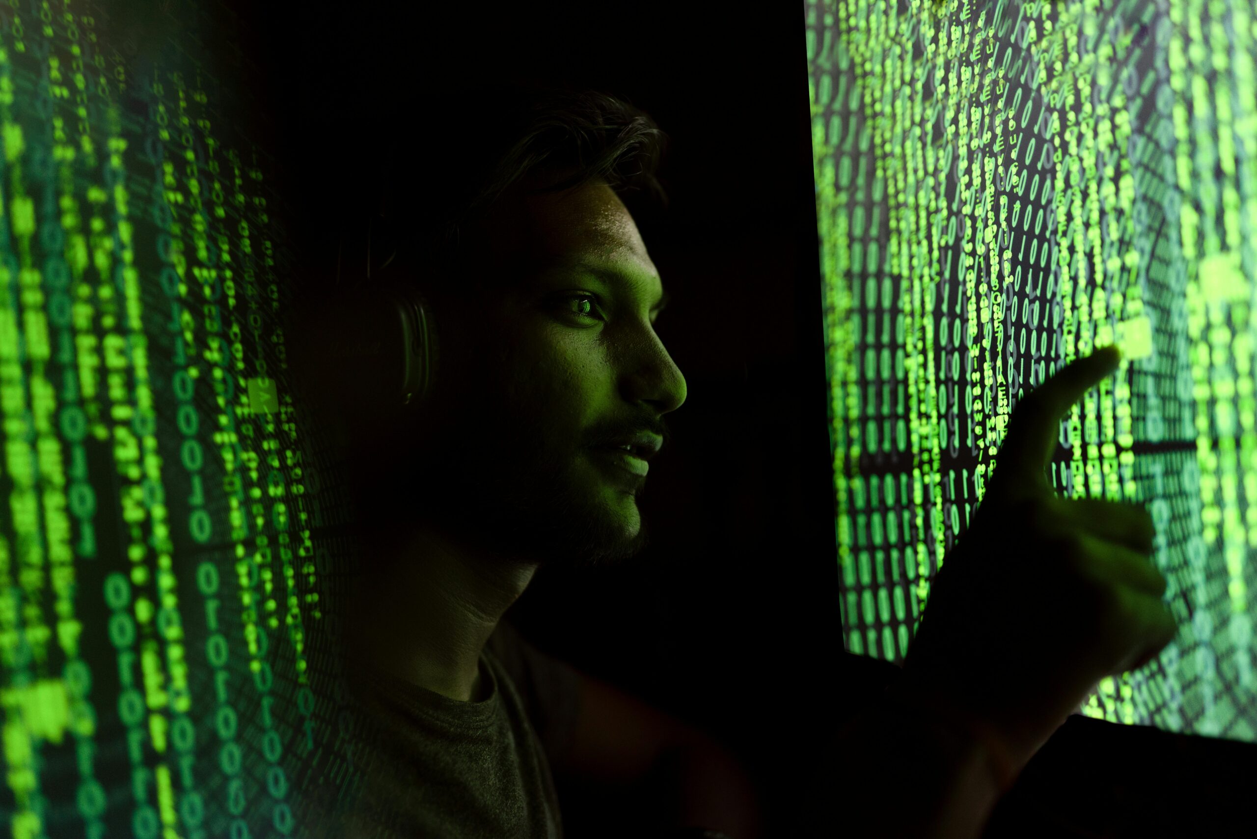 A man is stood in between two green neon text screens in a dark room. His face is lit green with an inquisitive look on his face as he stands facing one of the screens. His hand reaches out to touch one of the screens with his finger.