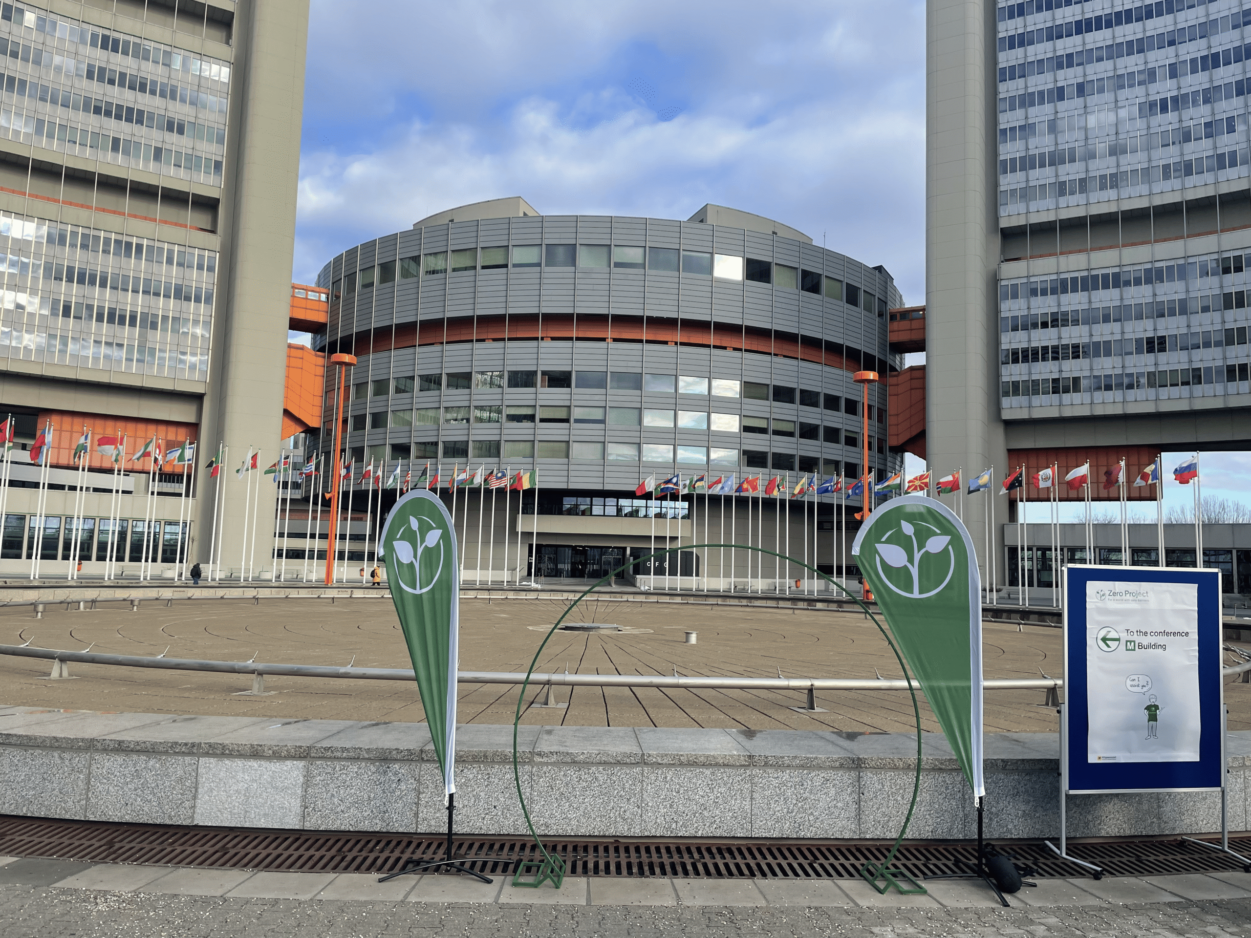 Exterior view of the United Nations complex in Vienna, showing the circular central conference building framed by two tall rectangular towers. Multiple national flags line the plaza in front of the entrance. In the foreground, two green Zero Project event banners with a white plant icon stand on either side of a circular green frame installation. To the right, a signboard reads “Zero Project – To the conference M Building,” with directional guidance. The sky is partly cloudy, and the open plaza space emphasizes the institutional scale of the venue and the international context of the event.