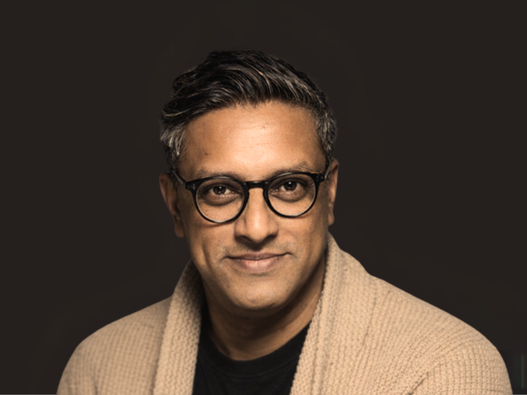 Professional headshot of Ravi Nath wearing round glasses and a beige textured coat over a dark shirt, looking directly at the camera with a calm and confident expression, set against a dark neutral studio background with soft lighting highlighting his face.